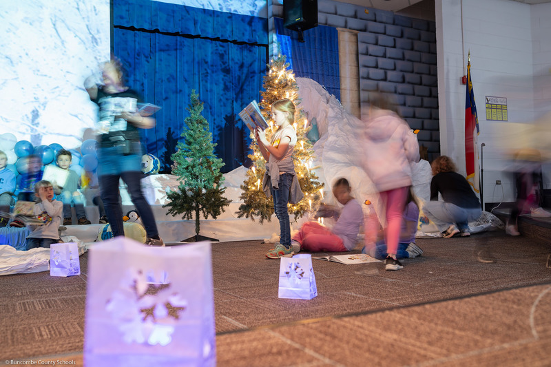 A student reads a book next to a lit Christmas tree while a long exposure causes blurry streaks as other people move through the scene..