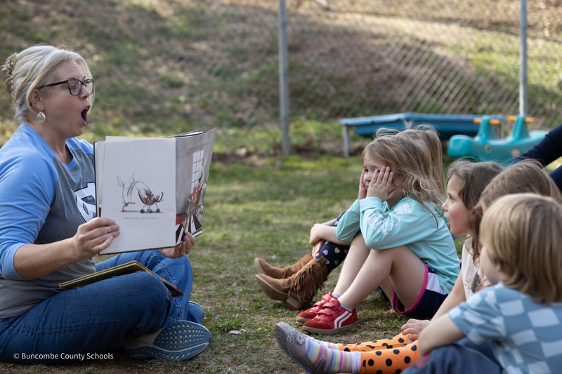Dr. Reed sits in the grass holding up a book to a group of preschool children.