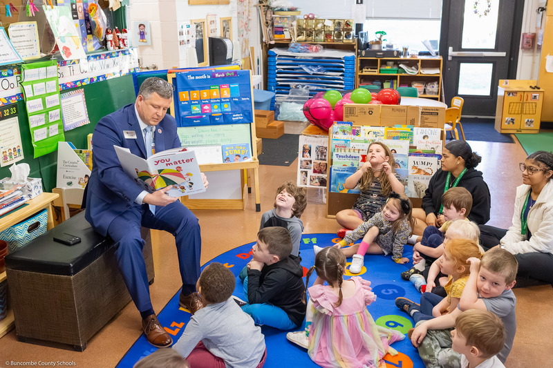 Mr. Cody sits on a bench with an open book. A group of preschoolers sit on a rug in front of him.