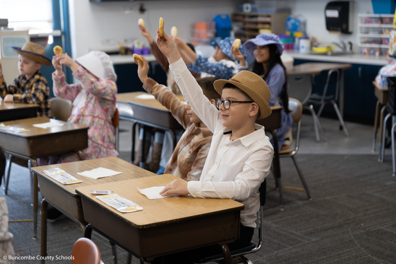 Classroom of students holding their Johnny cake in the air. 
