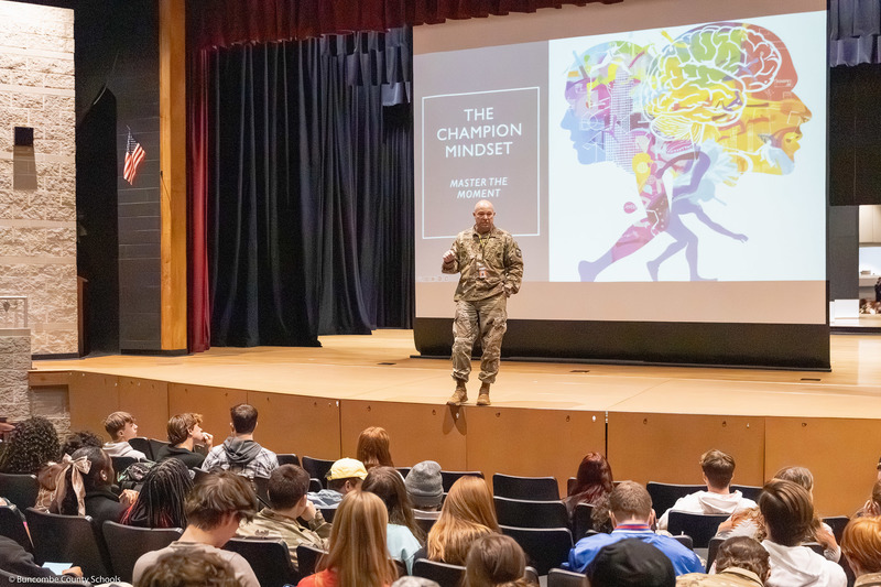 Chad Prevatte, JROTC instructor and track & field coach, speaks on stage with a presentation slide behind him.