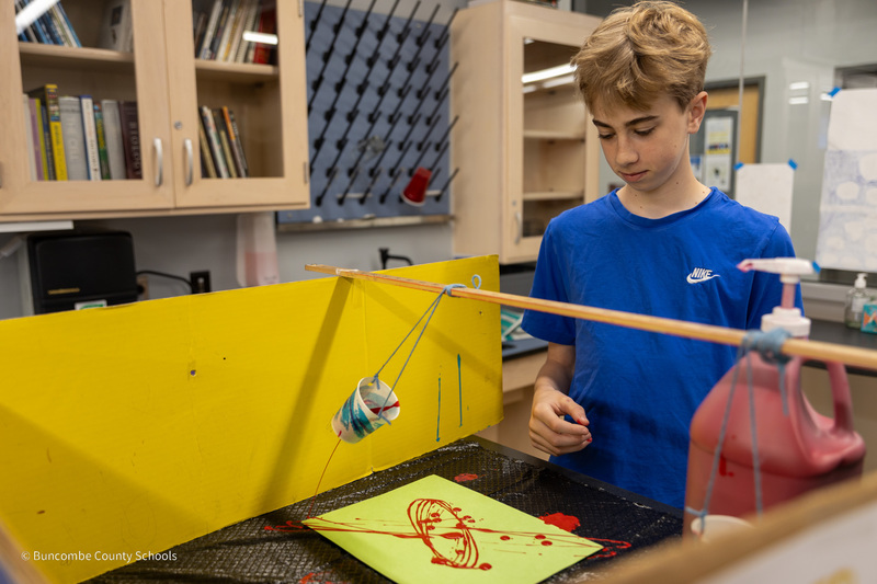 A student watches paint drip from the bottom of a cup as it swings and creates a piece of abstract art