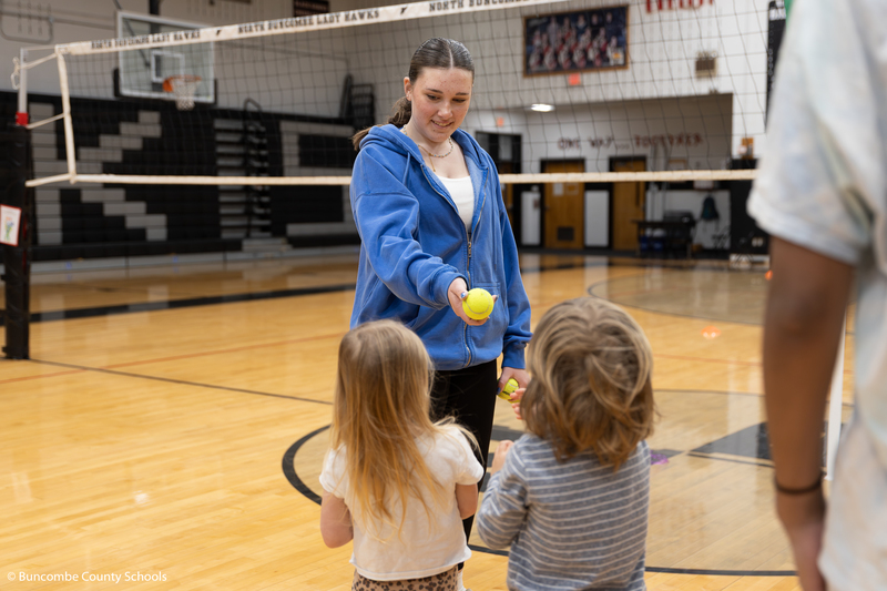 High school student handing a tennis ball to a preschooler. 
