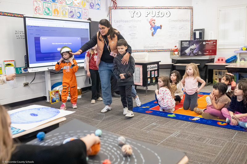 A teacher talks between two students, one of whom is wearing an astronaut costume.