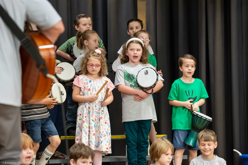 Students sing on stage while holding drums and rhythm sticks.