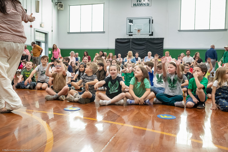 Students, seated on the gym floor, cheer and smile.