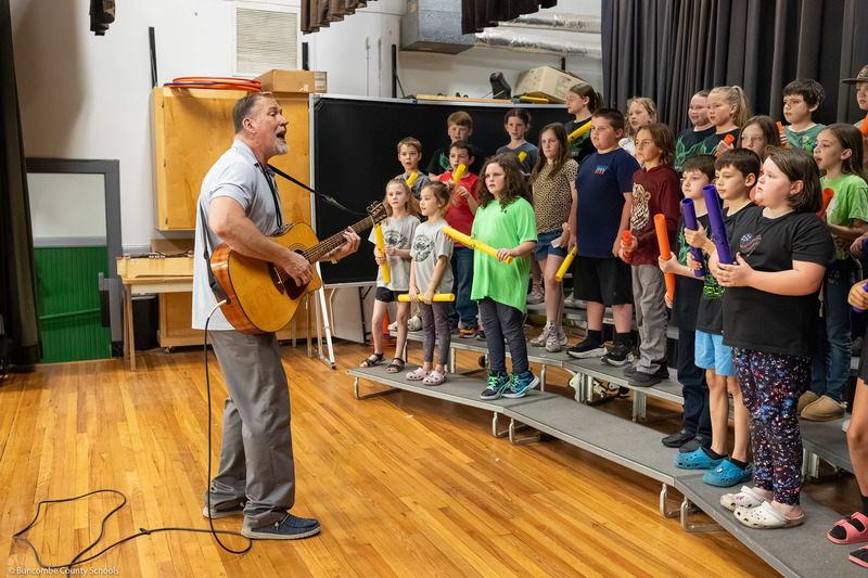 Barnardsville's music teacher leads a group of students in a song on stage with a guitar while the kids hold rhythm sticks.