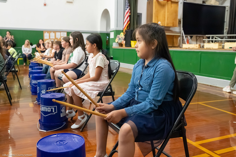 A student hits a bucket with drumsticks as part of a performance.