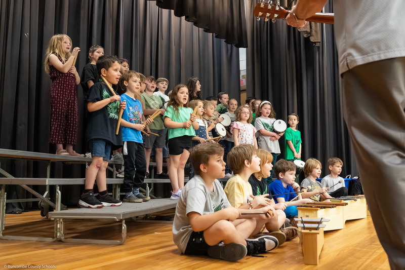 Students sing on stage while holding drums and rhythm sticks.