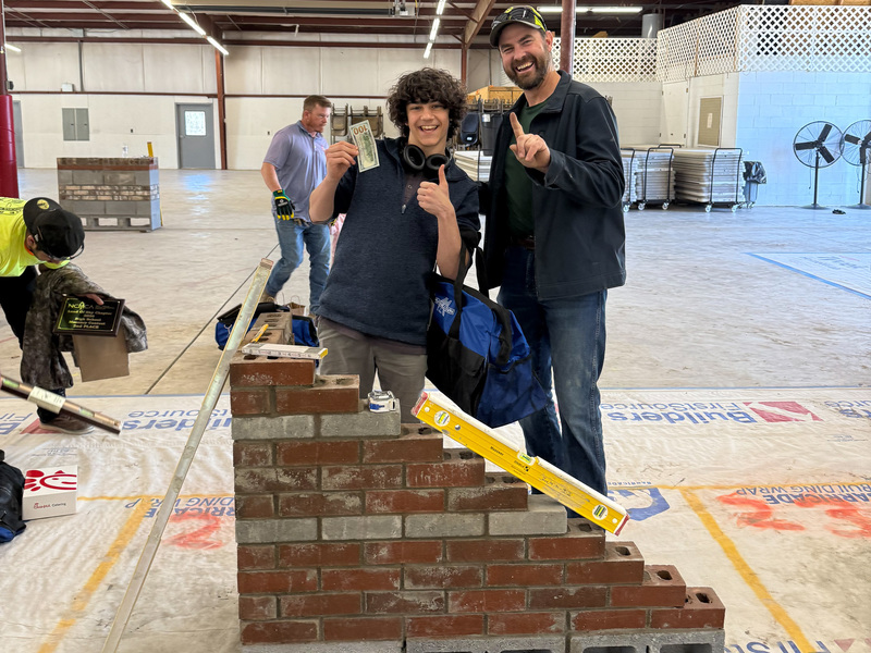 Mikhail holds his $100 prize in his hand as he stands next to Mr. Davis behind a wall he built for the competition.
