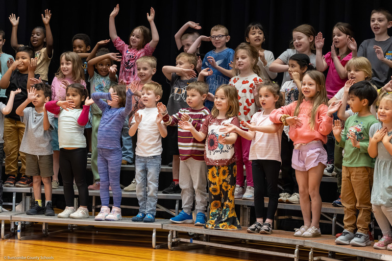 A group of students sing and wave their arms while standing on bleachers.