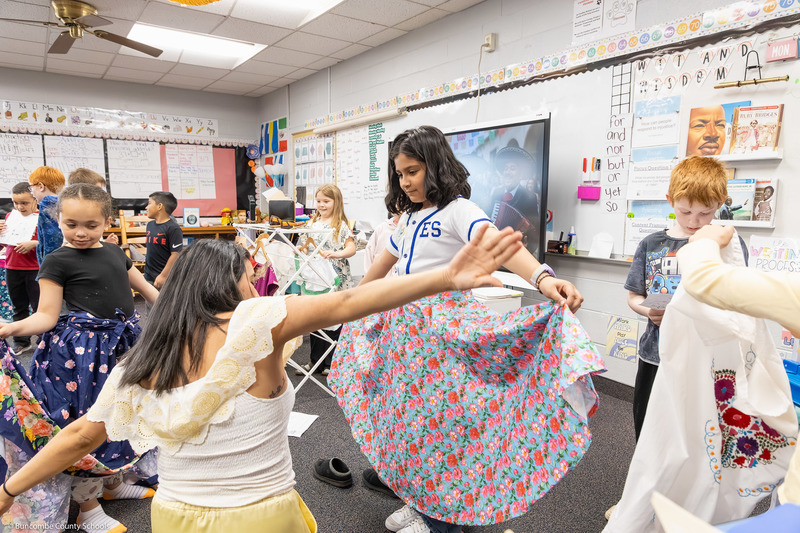 A girl tries on a cultural dress while her teacher demonstrates dance movements.