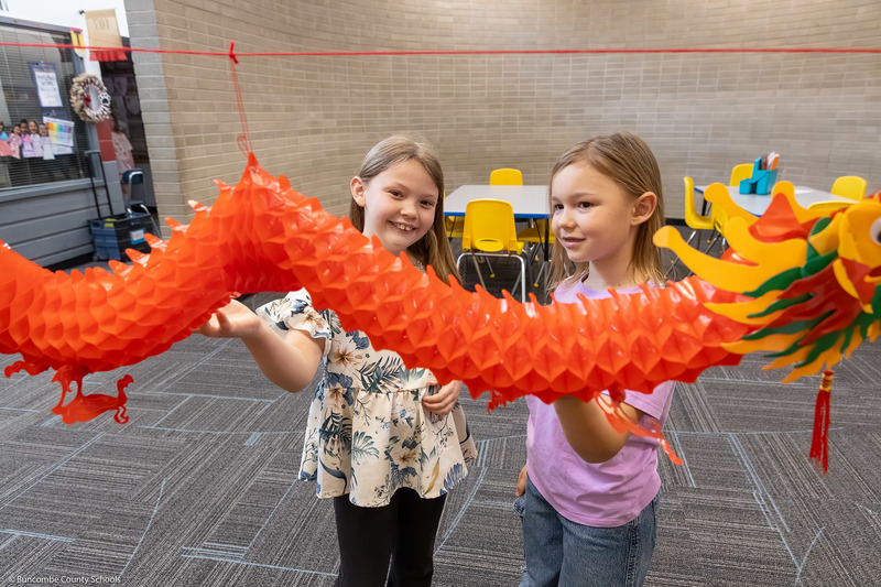 Two girls smile and point at a paper mache Chinese dragon.