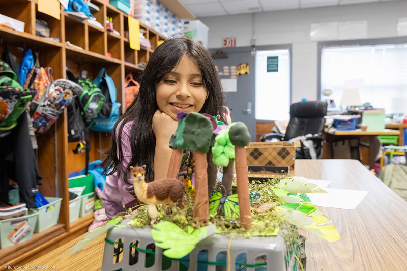 A girl leans on her elbows and smiles at a diorama showing a jungle scene.