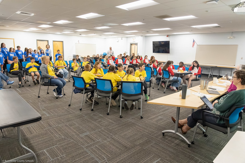 Teams sit at tables in a large conference room.