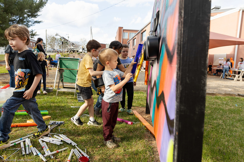 Students hold musical plastic tubes next to a music wall.