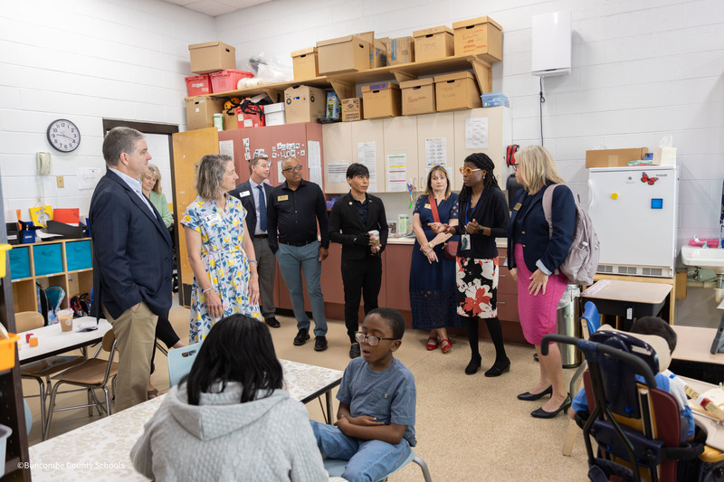 A student sits with his teacher in the foreground. Behind them, a group of lawmakers and school leaders listen to the principal speak.