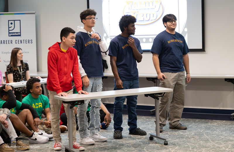 Four high school students stand behind a table participating in a Family Feud-style game while classmates watch from the side in a classroom setting.