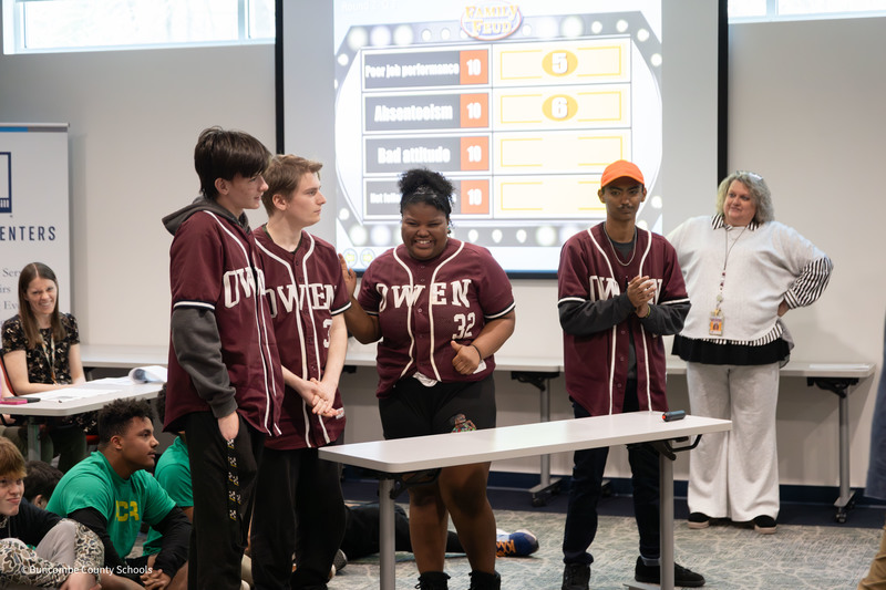 Four high school students in matching ‘Owen’ jerseys stand behind a table participating in a Family Feud-style game while classmates and staff watch in a classroom.