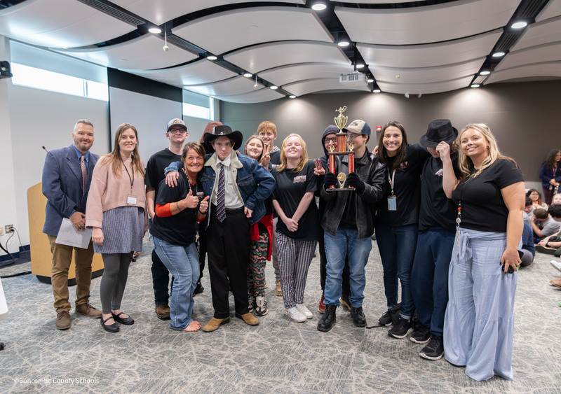 A group of high school students and staff pose together in a classroom, smiling and holding a trophy after a competition.