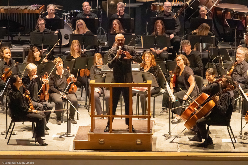 The conductor speaks to students from the stage with the orchestra behind him.