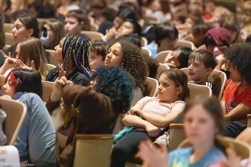 Students, sitting in rows,  listen to the performance.
