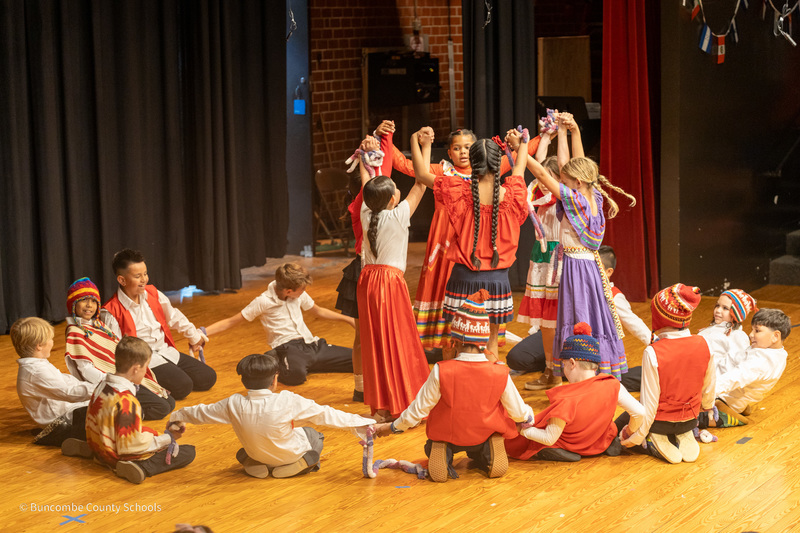 Girls stand on stage in a circle with locked arms as boys sit on the ground encircling the girls.
