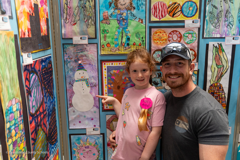 A student points to her snowman artwork while her dad kneels beside her.