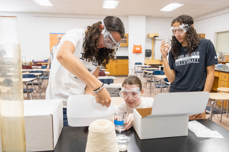 Three students, wearing safety goggles, pour water into a beaker.