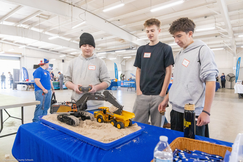A student operates a small remote control backhoe while two other students watch.