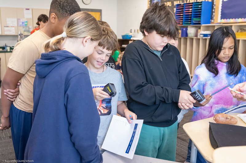 Students use an infrared thermometer to measure the temperatures of objects on a table.