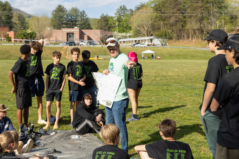 A Mills River Partnership representative points toward a group of students while holding a document.