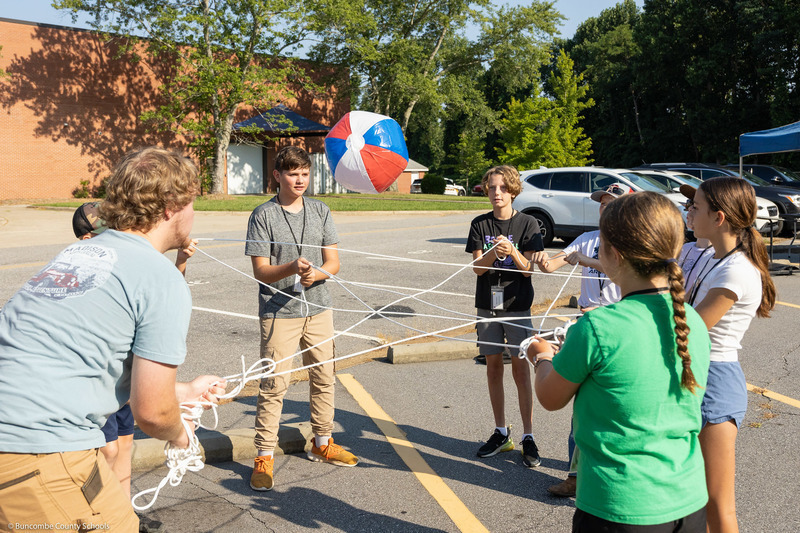 Students stand in a circle holding a net made of ropes and toss a beach ball.