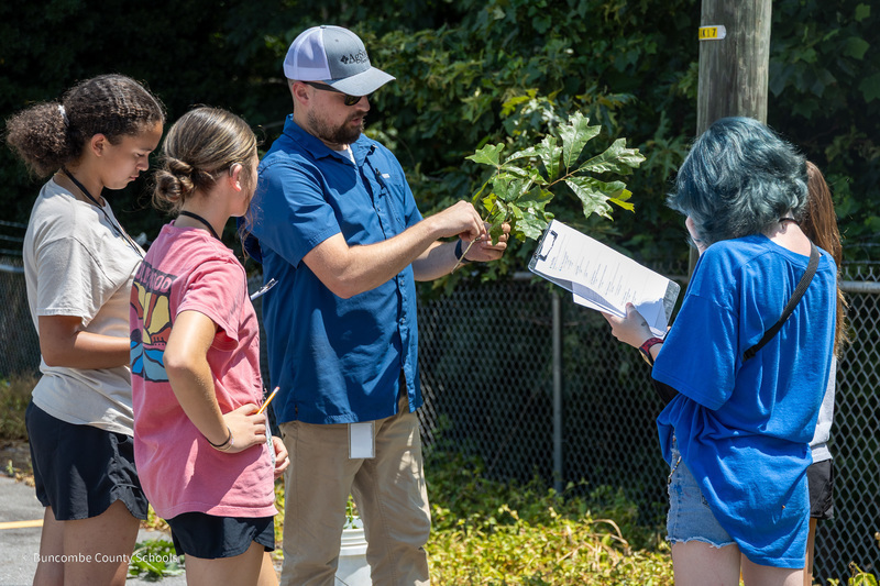 A teacher holds a branch while students with clipboards take notes.