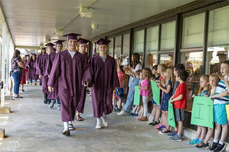 Graduates walking in a line down the breezeway with younger students lined up along the wall Graduates walking in a line down the breezeway with younger students lined up along the wall