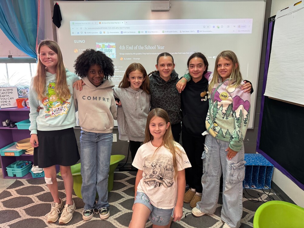 Seven elementary students pose together in a classroom after completing a songwriting project. Six students stand side by side with their arms around each other while one student kneels in front. Behind them, a projector screen displays the Suno music platform and a project titled “4th End of the School Year.” The classroom includes colorful bins, flexible seating, and a chart stand, creating a collaborative and creative learning environment.