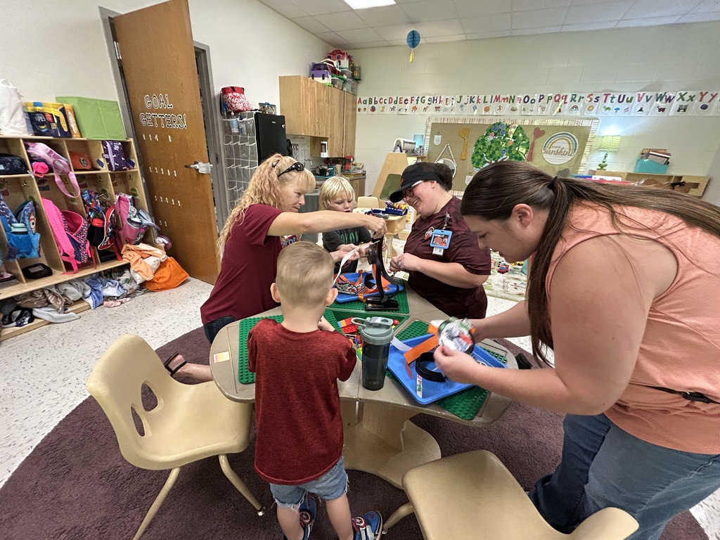 Alt text: A group of children and adults are gathered around a table in a preschool classroom, participating in an arts and crafts activity. One woman leans over to assist a child with their project, while others work on their own creations using blue trays filled with materials like ribbons and paper. The background features cubbies filled with backpacks and classroom supplies, as well as a wall decorated with an alphabet border and colorful artwork. The atmosphere is collaborative and focused, with everyone engaged in creative play.
