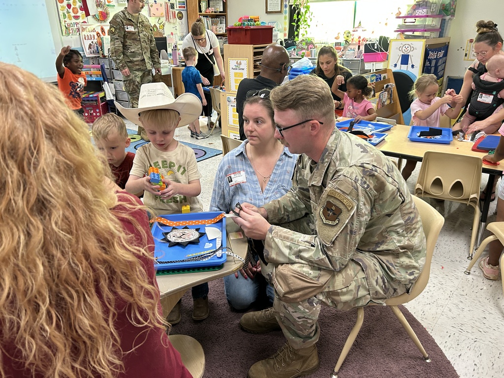 Alt text: A preschool classroom filled with children and adults engaged in hands-on activities. In the foreground, a young boy wearing a cowboy hat plays with colorful building blocks while sitting next to a woman and a man in military uniform who are interacting with him. A blue tray with a star-shaped badge and craft materials is on the table. Other children and adults can be seen working together at tables in the background. The classroom is vibrant, decorated with educational materials, artwork, and organized learning centers. The atmosphere is warm, busy, and collaborative.