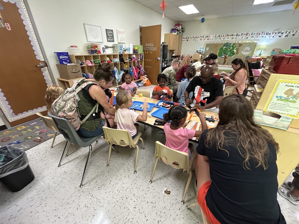 Alt text: A busy and colorful preschool classroom where children and adults sit around tables engaging in arts and crafts activities. The children are using blue trays filled with craft materials, while adults assist and interact with them. The classroom is decorated with educational posters, cubbies filled with supplies, and alphabet letters across the top of the wall. A sign labeled "Puzzle Center" is visible in the foreground, and the atmosphere appears lively and collaborative.