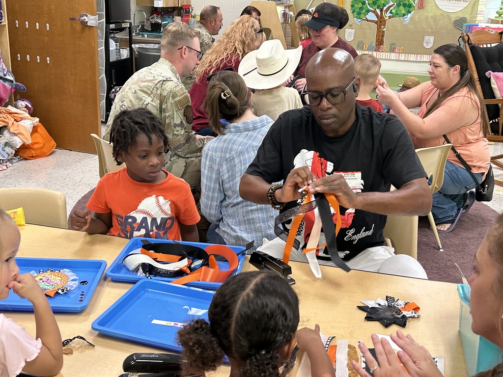 Alt text: A man wearing glasses and a black graphic t-shirt helps children with an arts and crafts project at a classroom table. He is focused on tying orange, black, and white ribbons together. Around him, several children work on similar projects using blue trays filled with craft supplies. In the background, more adults and children, including individuals in military uniforms, are engaged in creative activities. The classroom is colorful and filled with educational decorations, fostering a collaborative and hands-on learning environment.