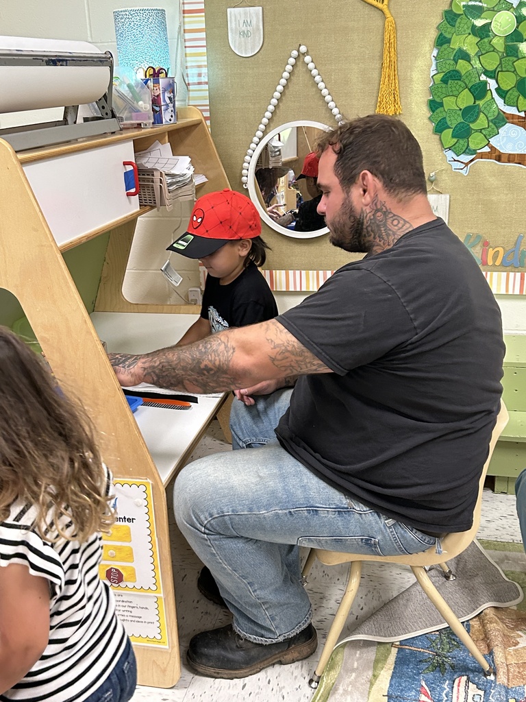 Alt text: A man with tattoos sits beside a young child wearing a red Spider-Man cap at a small wooden workstation in a classroom. They appear to be working together on a project, with craft supplies like paper and scissors nearby. The classroom wall behind them features a round mirror, decorative elements, and a tree-shaped bulletin board labeled “Kind.” A young girl in a striped shirt stands in the foreground, slightly out of focus. The scene conveys a warm, collaborative learning environment.