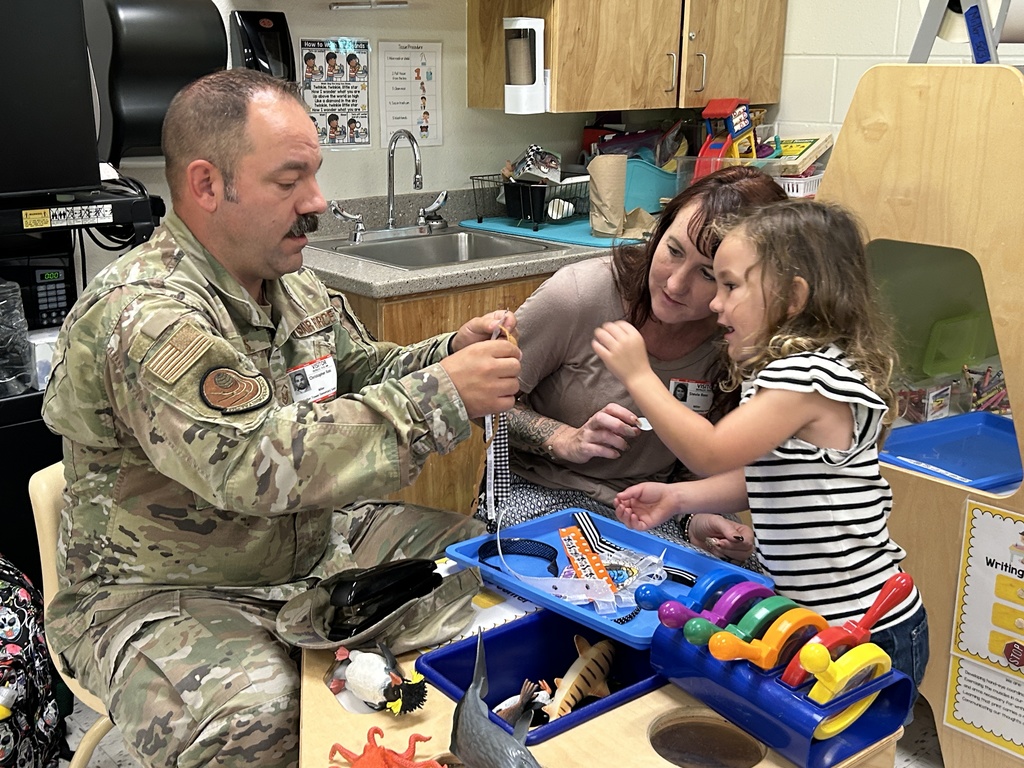 Alt text: A man in a military uniform, a woman, and a young girl are seated at a small table in a classroom, engaging in a hands-on activity. The man is holding up a piece of ribbon while the girl reaches toward it excitedly, and the woman smiles as she watches. Various craft supplies and toys are scattered on the table, including a toy tiger, plastic insects, and a colorful rolling toy. The background shows a classroom sink and cabinets, with educational materials and posters on display. The scene captures a moment of creativity, learning, and connection.