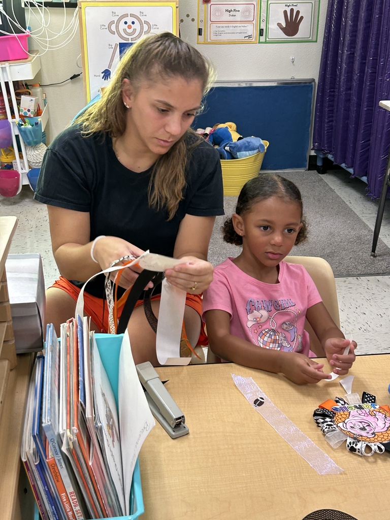 Alt text: A woman and a young girl are seated at a classroom table, working together on a craft project involving ribbons. The woman holds several strips of ribbon in her hands, while the girl looks on and helps. The table is covered with craft materials, including a finished decorative ribbon project featuring colorful loops and a cartoon character. Behind them, classroom decor includes educational posters and a bin of stuffed animals, contributing to a warm and engaging learning environment.