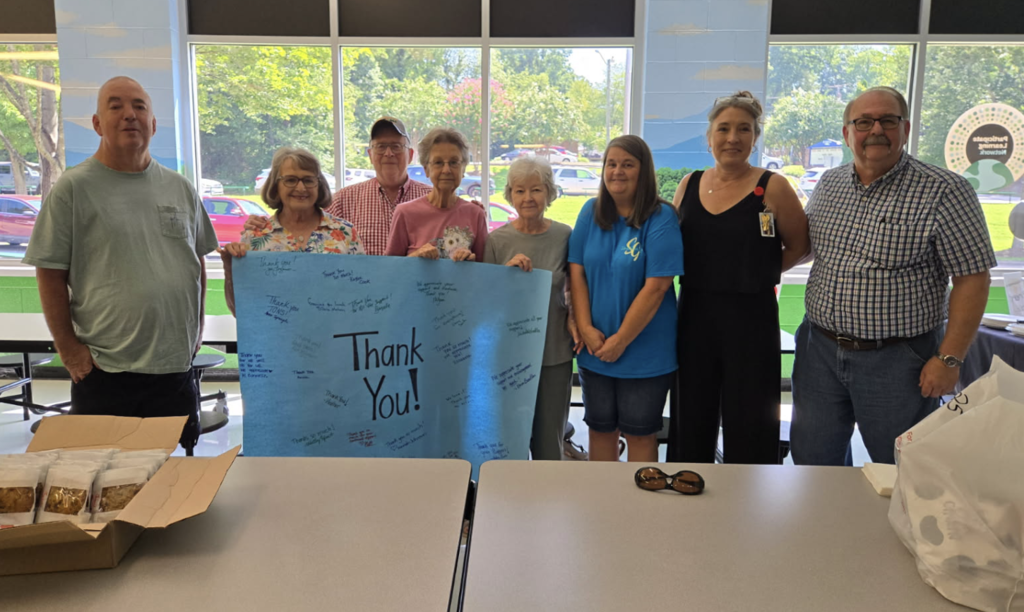 Seven adults stand together indoors, holding a large blue “Thank You!” poster covered with handwritten messages. Tables with food containers and personal items are in the foreground, and large windows behind the group show trees and a parking area outside.