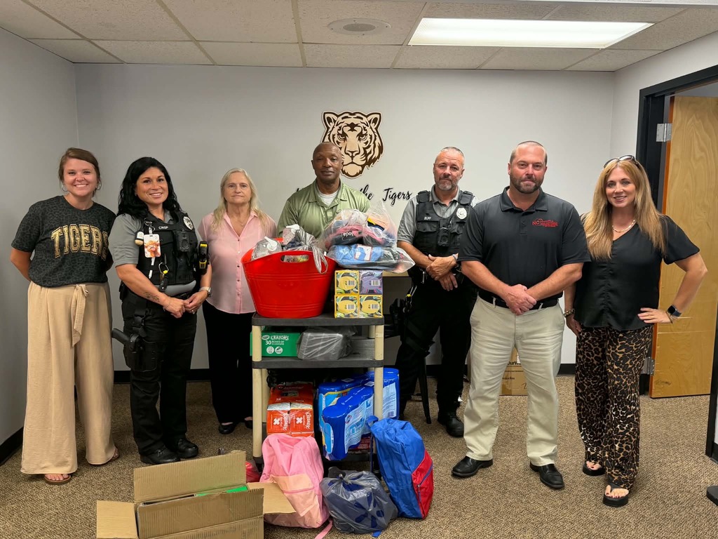Seven adults stand together inside Valdese Elementary beside a cart filled with donated school supplies, including backpacks, crayons, tissues, and other classroom items. A tiger logo is displayed on the wall behind the group.