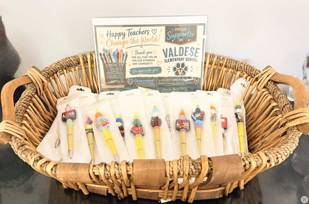 A large woven basket filled with individually packaged, colorful teacher-themed pens donated to Valdese Elementary School. Behind the basket is a sign from Grounded Home Inspections that reads, “Happy Teachers Change the World!” and thanks Valdese teachers for all they do for students and the community.