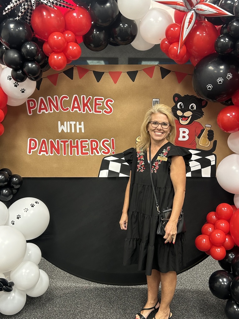 Woman in black dress standing in front of colorful  balloon arrangement and banner saying Pancakes with Panthers!