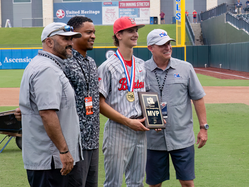man in a gray hat and matching gray shirt standing next to a man in a black camo shirt with an ID around his neck standing next to a high school boy in a baseball uniform and medal holding an MVP award, next to another man wearing the same hat and shirt as the first man.  All are lined up on the baseball field smiling at another camera.