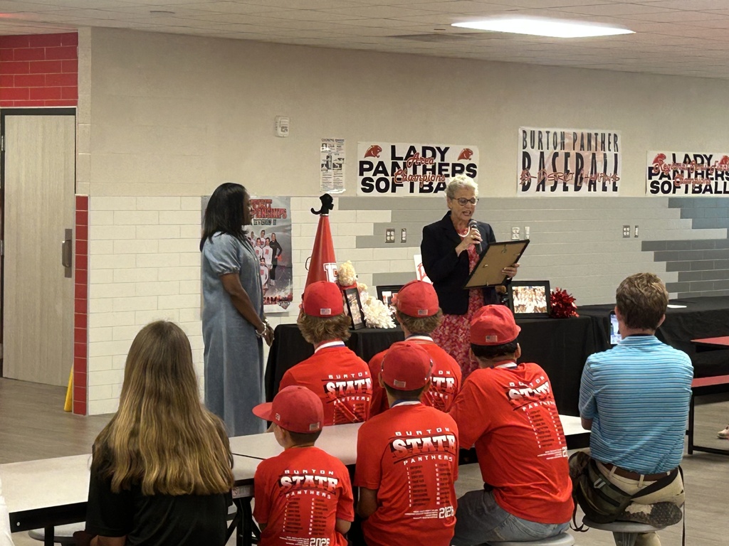 a lady talks into a mic while holding a frame while another woman stands watching her and 7 people sit in front of them watching.