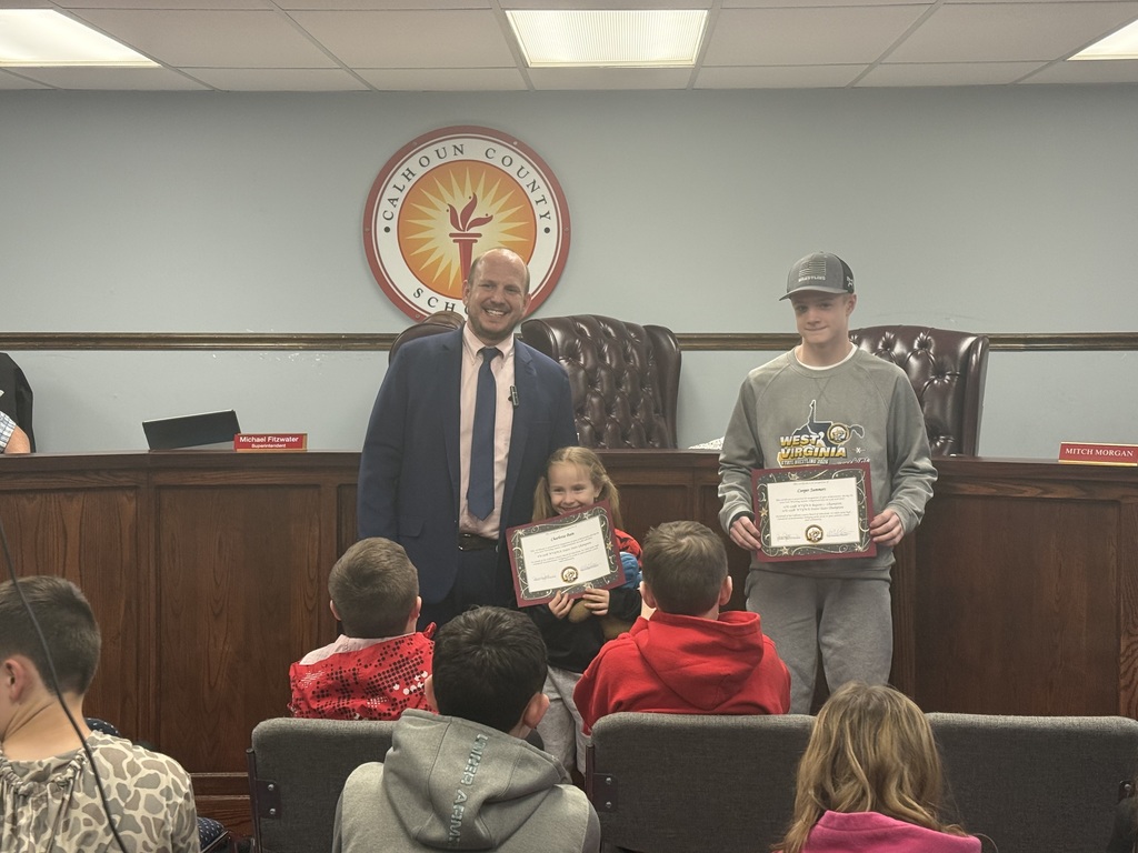 A group is gathered in a school board meeting room. Two students are standing at the front holding certificates: one student holding an award for Cooper Summers, 12U 125lbs Class; State Champion and Region 1 Champion, and a younger student holding an award for Charlotte Bain, 6U 50lbs Class; State Champion. An adult stands beside them near the board’s wooden desk, while several other students sit in chairs facing the front. The Calhoun County Schools emblem is displayed on the wall behind them.